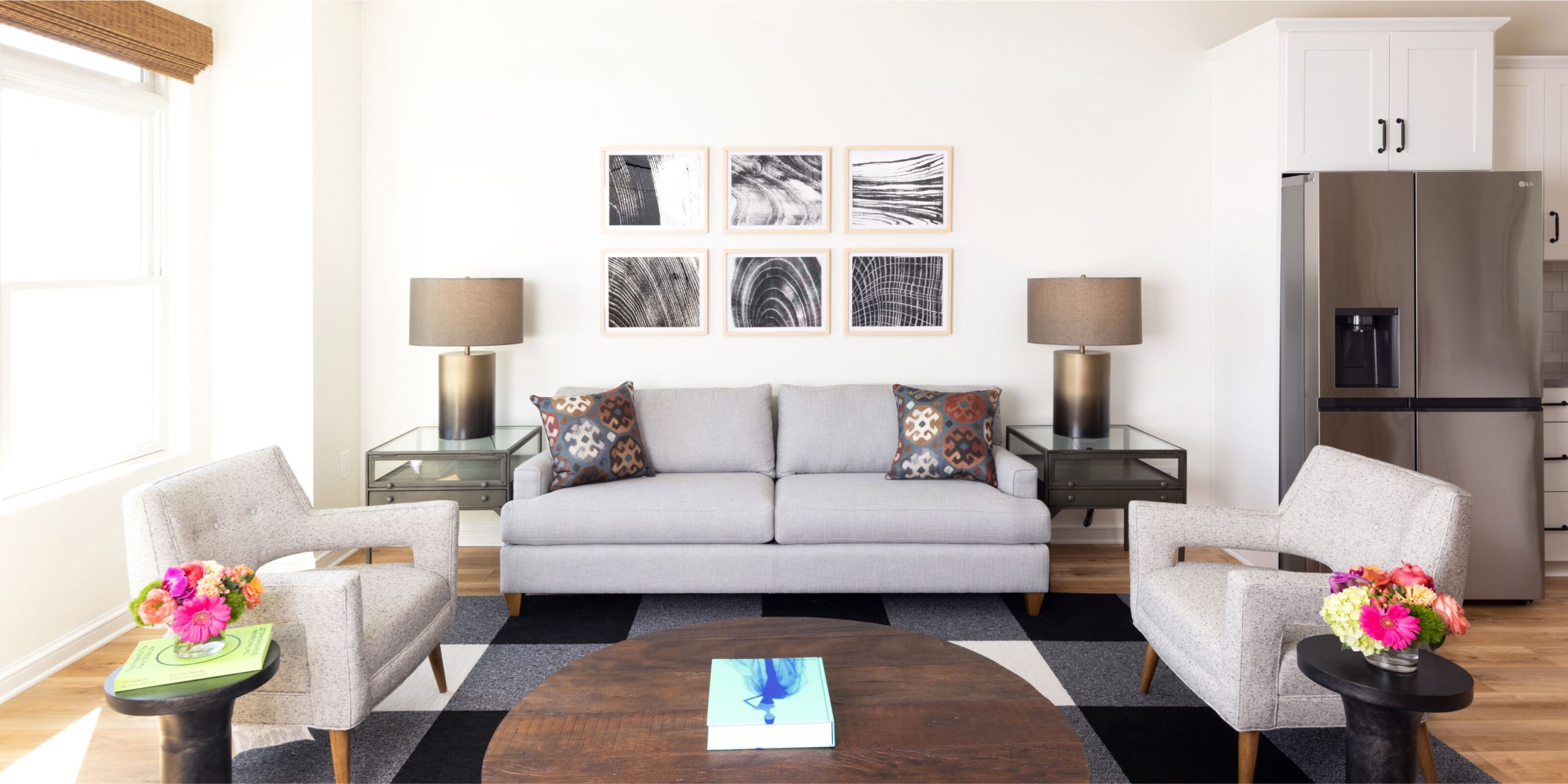 A stylish living room featuring a grey sofa with patterned throw pillows, two light grey armchairs, and a round wooden coffee table on a black and white checkered rug. The walls are adorned with black and white framed art, and there are two modern table lamps on either side of the sofa.