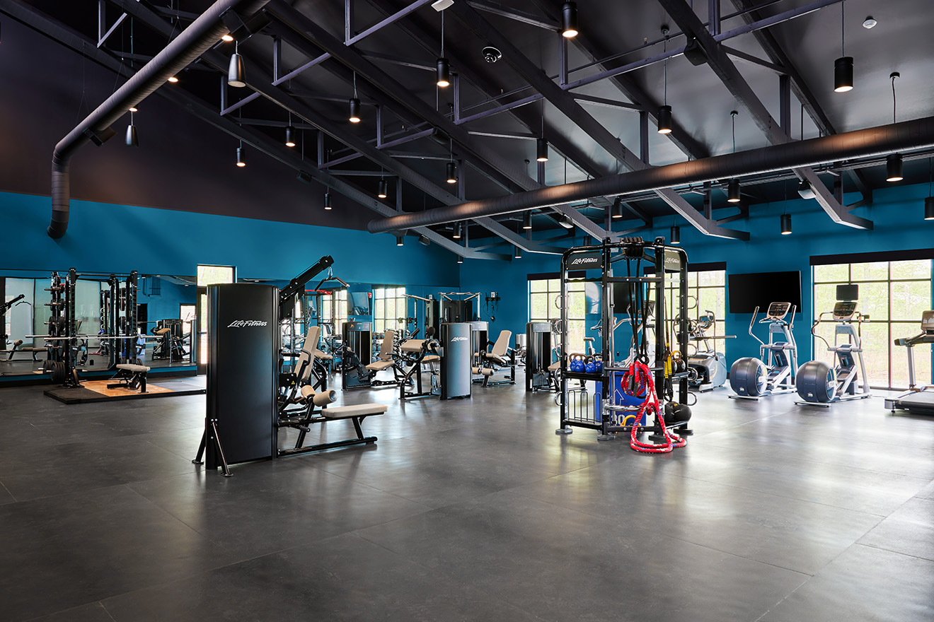 A spacious fitness center with various exercise equipment including weight machines, treadmills, and stationary bikes. The room has a high ceiling with exposed beams and large windows allowing natural light to fill the space