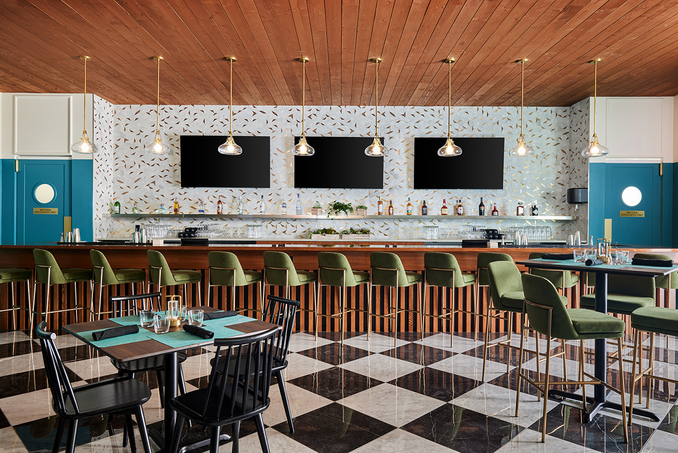 A contemporary bar and dining area featuring a long wooden bar with green upholstered bar stools, black and white checkered floor, and a terrazzo backsplash with three large TVs mounted above the bar. Modern pendant lights hang from the ceiling, and there are tables and chairs for additional seating